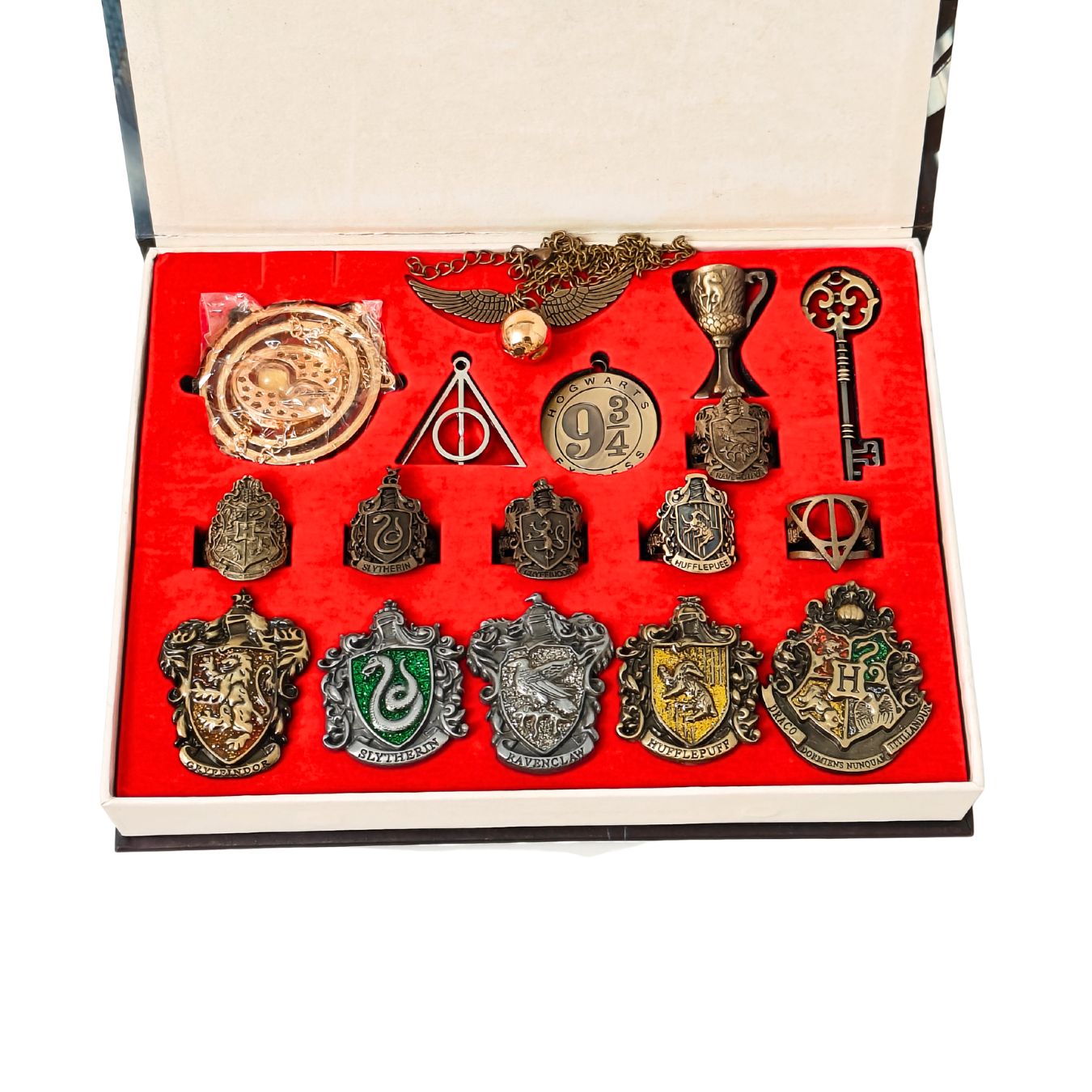 A set of Harry Potter themed jewelry and accessories displayed inside an open presentation box with a red velvet insert. The collection includes a Deathly Hallows pendant, a Sorting Hat, a key, a cup, and multiple Slytherin, Ravenclaw, Gryffindor, and Hufflepuff crest pins.